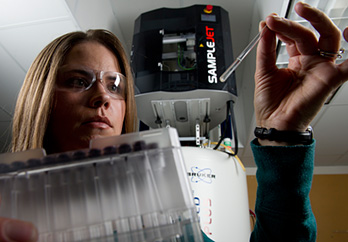 Photo of a woman wearing safety glasses in a laboratory looking at pipette samples and holding box of test tubes