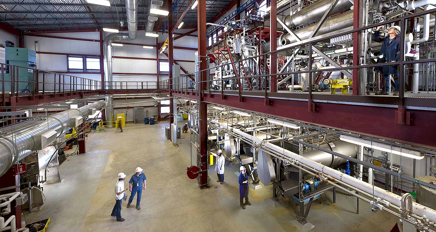 Photo of the interior of an industrial, two-story building with high-bay, piping, and large processing equipment. Three workers are in hard hats.
