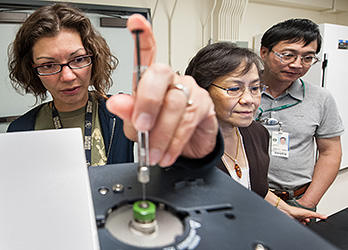 Photo of two women and one man in a laboratory measuring hydrogen production of engineered microbes in a gas chromatograph.