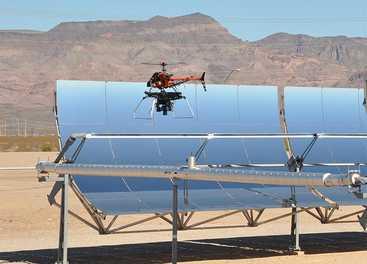 A small helicopter drone hovers near the top of a parabolic trough in the desert in front of mountains.