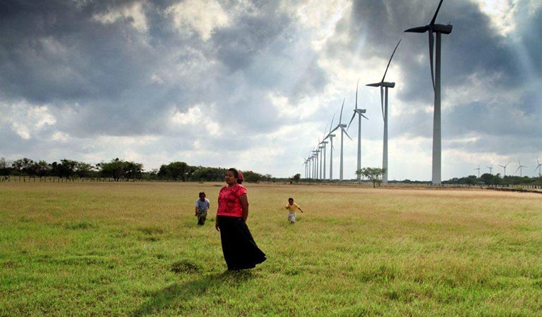 Photo of a Mexican woman and two children in a field with large wind turbines behind them.