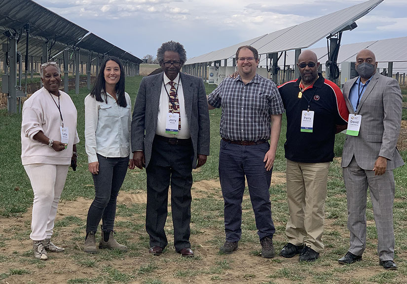 Six people stand in front of commercial-scale solar panels on a farm.