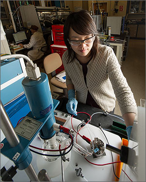 In this photo, a woman in safety glasses works in front of a device that includes plastic tubing and red and black wires.