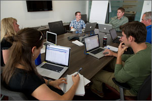 In this photo, six NREL employees sit around a large brown table, all with their laptops open.