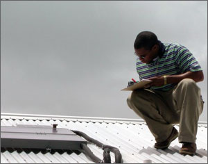 This photo shows a young man on an aluminum rooftop crouching as he looks at a solar collector and writes some data on a clipboard.