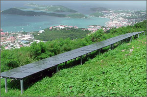 In this photo, a long rectangular chain of solar panels sits in the foreground, surrounded by lush green vegetation. In the background is the Caribbean Sea dotted with large rocks.