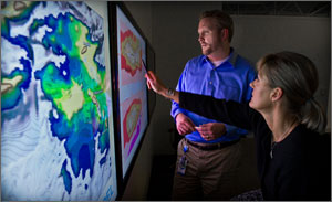 In this photo, a man and a woman are looking at a huge colorful wall map that shows sun resources in yellows and oranges, wind resources in blues.
