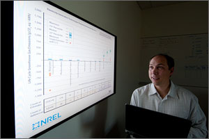 In this photo, a scientist holds a laptop, looking up at a projection that graphs greenhouse-gas emissions for various generation technologies.  