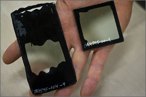 This is a close-up of a square and a rectangular sample held in a hand. The rectangular sample shows failure in the form of black globs invading the transparent center.