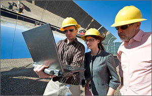 This photo shows three people in yellow hard hats looking at a laptop computer screen, with parabolic mirrors in the background.