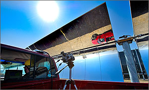 In this photo, a red pickup rumbles alongside a row of parabolic-shaped mirrors while the upside-down reflection of the truck is visible along the upper sections of the mirror.