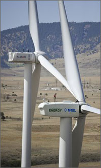 Photo of a giant wind turbine, with a man standing on the turbine's nacelle.