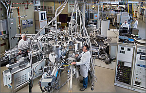 This photo shows two men in white lab coats amid gleaming silver machinery with hoses heading to an unseen ceiling. 