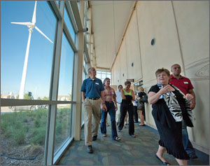 In this photo, a woman leads a half dozen others down a corridor lined with glass windows. Through the windows, a large, white wind turbine can be seen.
