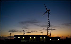 This photo is in twilight, with the setting sun contrasting with the dark clouds. In the foreground is a low-rise hotel, with wind turbines behind.