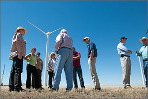 In this photo, seven people listen to a man with his back to the camera. In the background are a wind turbine and a brilliant blue sky.