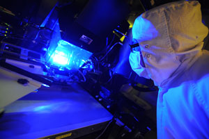 This photo shows a photolithography tool emitting blue light from its core. A man in full sterile uniform, including face mask, is looking at the tool.