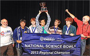 Two men and five teenage boys stand behind a banner that reads “U.S. Department of Energy Office of Science National Science Bowl 2013 Regional Champion.” The boy in the center holds a medal over his head.