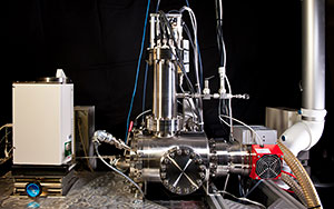 Photo of a complex instrument comprised of a stainless-steel cylinder aligned horizontally and a smaller stainless-steel cylinder rising vertically. To the left is a plastic-lined box.