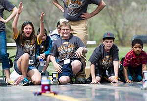 In this photo, four middle schoolers are crouched at the edge of a race track. A girl on the left holds up both hands signaling victory, while the boy next to her smiles. To the right are two boys whose faces register concern as they watch their car move down the track.