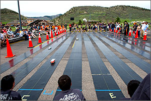 In this photo, four cars can be seen on the neoprene tracks, bordered by orange construction cones. In the background are several middle-school students and behind them, the foothills.