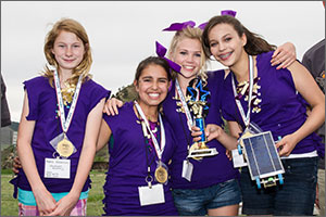 In this photo, four girls in purple tops and purple hair ribbons smile at the camera. The one on the right holds a trophy and the solar-powered model car. All four girls are wearing medals around their necks.
