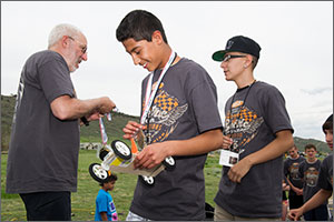 In this photo, a man in a white beard and glasses presents a trophy to a middle-school boy who is holding a car made of a sheet of balsa wood. A second boy awaits his turn to get a medal. All three are wearing the official electric car competitions grey T-shirt.