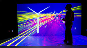 In this photo, a man in silhouette holding a remote-control device stands in front of a giant screen looking at ribbons of yellow flowing past a wind turbine while purples, whites, and blues enhance the three-dimensional effect.
