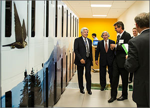 In this photo, three men share a laugh as they stand in front of a high-performance computer (HPC) that takes up the entire area to their right. The HPC has a mural of trees, mountains, and a bird of prey.