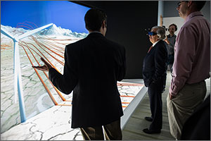 In this photo, a man wearing high-tech glasses gazes at a huge screen displaying a wind turbine. Ribbons of orange represent the way wind flows downstream when the blades hit it. A man with his back to the camera explains the process, while two others look on.