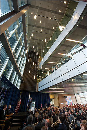 This photo shows a seated crowd watching a man in a suit at a raised podium. The upper two-thirds of the photo shows windows, hanging pendant lights, and breezeways.