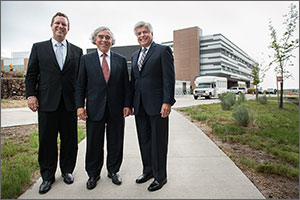 In this photo, three men in dark suits stand on a sidewalk smiling. Behind them is a three-story laboratory building, with a shuttle bus parked outside.
