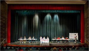 Photo of a stage in an auditorium; onstage are two teams sitting at tables to compete.