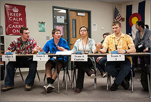 Photo of four high school students sitting at desks and holding hands and concentrating.