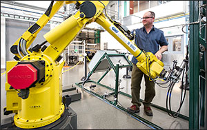 In this photo, a man stands behind a large yellow robot with his hands on the robot's arm. 