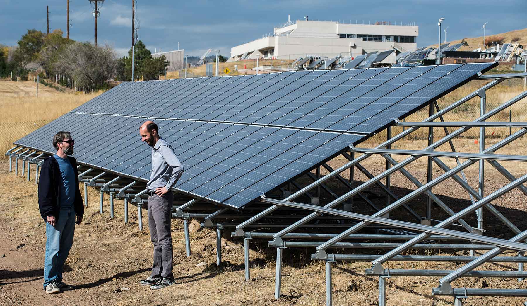 Two men stand next to solar modules in a field on the NREL campus.