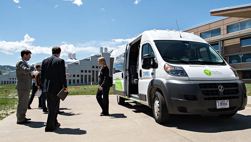 On a business campus, a woman exits a white van with an open door as people stand nearby.