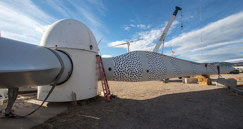 A turbine rotor sits perpendicular to the ground. One of the two visible blades has strips with dots along the length of it. A crane, two other turbines, and two meteorological towers are in the background.