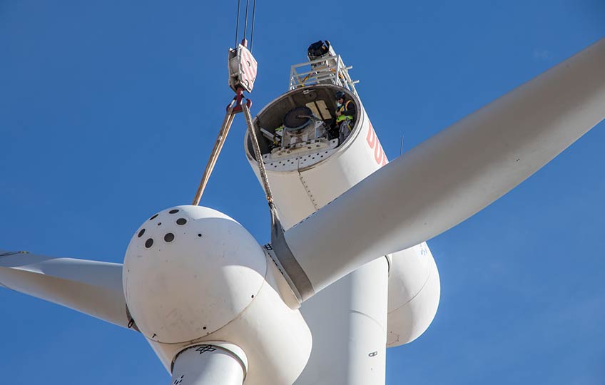A three-bladed rotor hub is suspended from straps on a crane hook several feet below the turbine nose. A worker in safety vest, helmet, and safety glasses watches from the open nacelle.