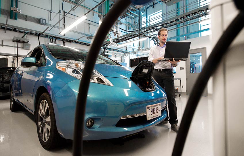 Man standing in lab garage looking at his laptop, standing next to an electric vehicle with charging cables in the foreground.