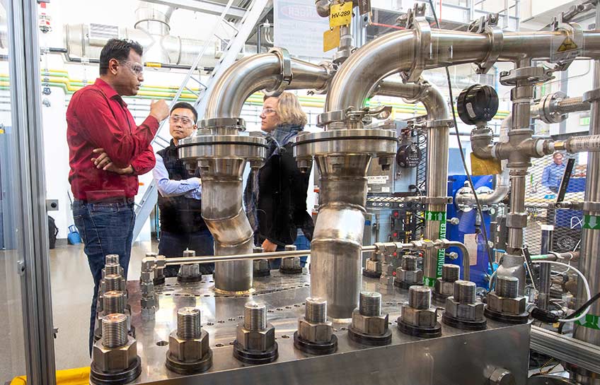 Photo of a large piece of machinery with silver pipes that produces hydrogen. People with protective goggles stand in the background discussing the equipment.