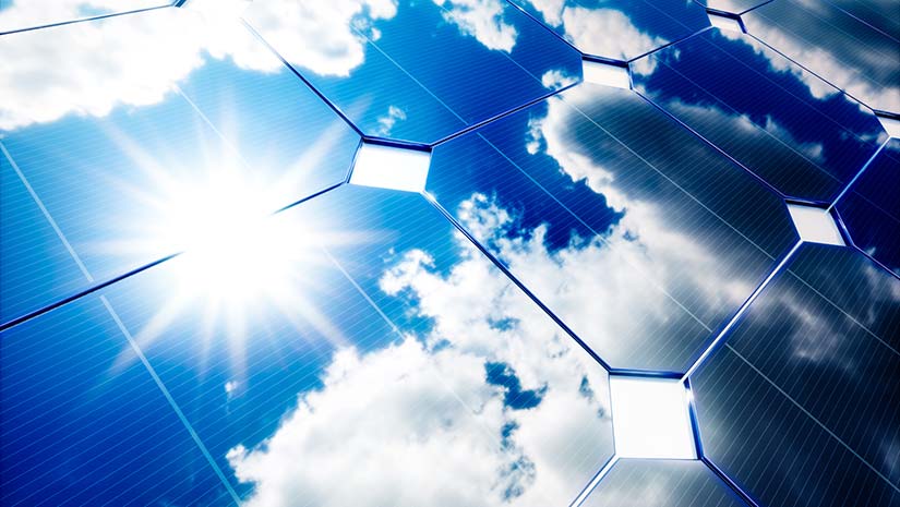 Photo of a close-up view of a solar panel with sun and blue skies in the reflection. 
