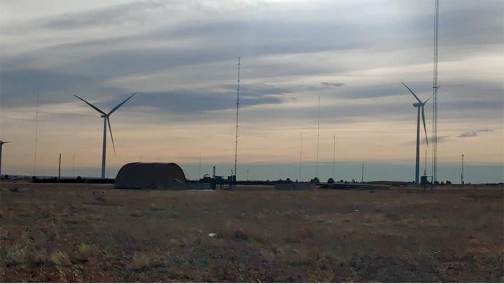 An open landscape at twilight with wind turbines and a Quonset hut in the distance