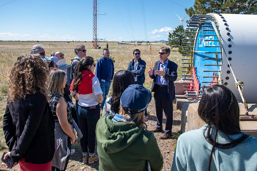 A group of students listen to a man standing next to a wind turbine blade laying horizontally on a platform. The turbine is stamped with NREL’s logo.