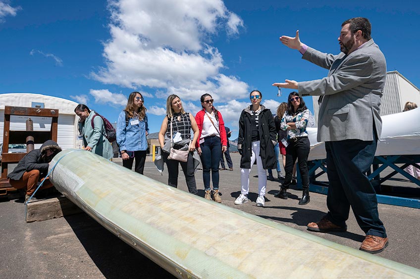 A man stands with his arms extended demonstrating a concept to a group of people listening to him. They all stand behind a wind turbine blade laid on the ground.