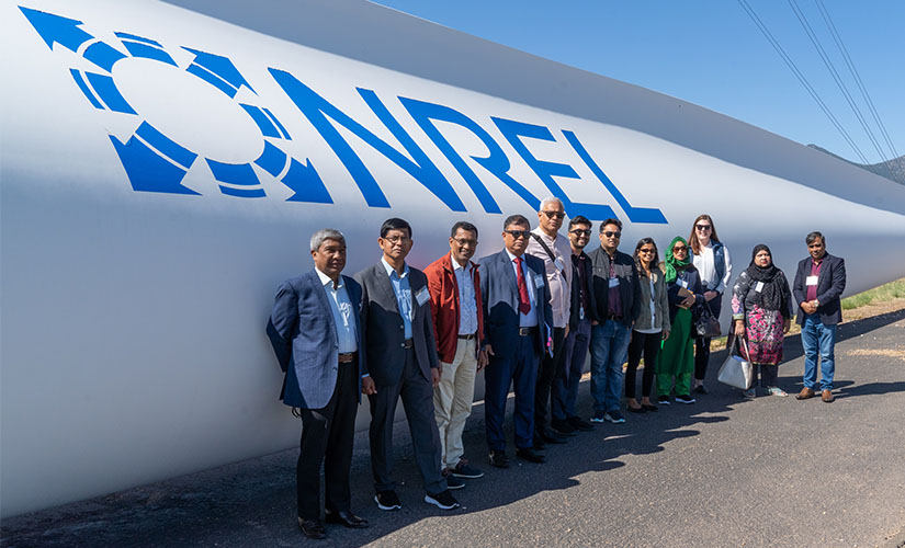 On a sunny day, the group of nine smiles for a photo in front of a turbine blade laying on the ground with the NREL logo on the side.