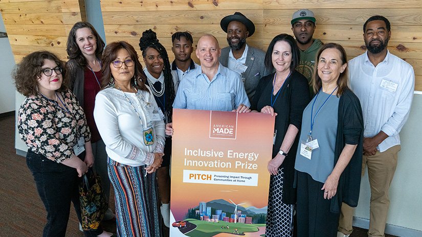 Eleven people stand smiling in front of a poster for the Inclusive Energy Innovation Prize PITCH event.