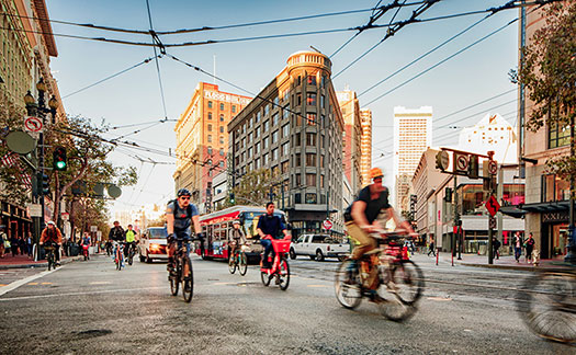 Photo of people riding bicycles on San Francisco city street, with bus and cars in background, cable car wires above, and pedestrians on sidewalks.