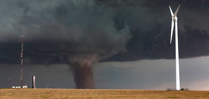 A wind turbine stands in a flat field with a tornado in the background.