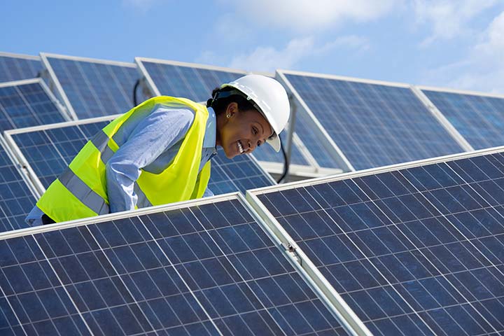 Female in a yellow safety vest and hard hat smiling next to solar photovoltaic panels.